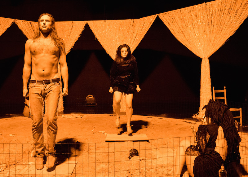A bare chested man in jeans and long hair stands at the edge of a sand-filld stage. Another performer sits with her back again wire fencing and Ann Liv Young jumps upstage in a black dress